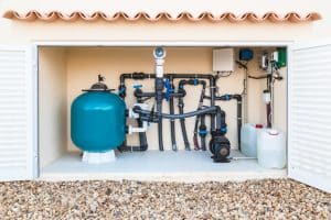 Swimming pool equipment system with a large filter tank, pump, connected pipes, valves, and chemical containers inside an outdoor equipment enclosure.
